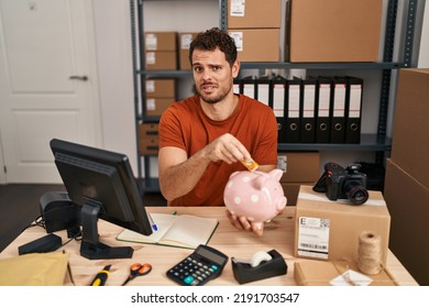 Young Hispanic Man Working At Small Business Ecommerce Holding Piggy Bank Clueless And Confused Expression. Doubt Concept. 