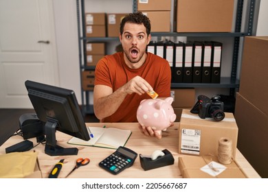 Young Hispanic Man Working At Small Business Ecommerce Holding Piggy Bank Afraid And Shocked With Surprise And Amazed Expression, Fear And Excited Face. 