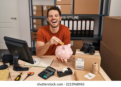 Young Hispanic Man Working At Small Business Ecommerce Holding Piggy Bank Smiling And Laughing Hard Out Loud Because Funny Crazy Joke. 