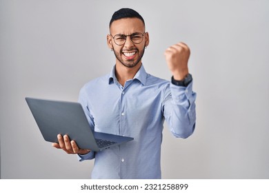 Young hispanic man working using computer laptop angry and mad raising fist frustrated and furious while shouting with anger. rage and aggressive concept. 