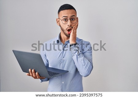 Young hispanic man working using computer laptop afraid and shocked, surprise and amazed expression with hands on face 