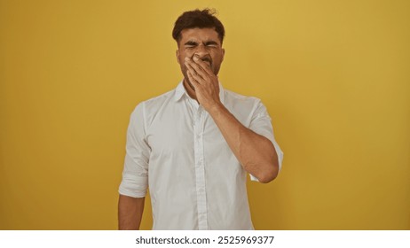 Young hispanic man yawning while covering his mouth, standing over an isolated yellow background. - Powered by Shutterstock