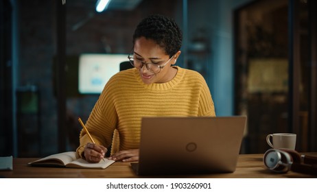 Young Hispanic Marketing Specialist Working On Laptop Computer In Busy Creative Office Environment. Beautiful Diverse Multiethnic Female Project Manager Is Writing Down Notes In Paper Notebook.