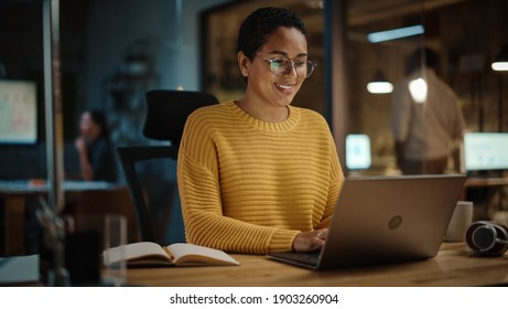 Young Hispanic Marketing Specialist Working on Laptop Computer in Busy Creative Office Environment in the Evening. Beautiful Diverse Multiethnic Female Project Manager Smiling.