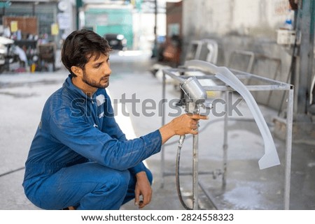 A young Hispanic mechanic squats while pointing a spray gun at a car bumper in an auto repair shop.