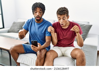 Young hispanic men using smartphone sitting on the sofa at home pointing down with fingers showing advertisement, surprised face and open mouth