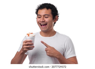 A young Hispanic, Mexican, eats corn in a cup, esquite, with white cheese and spicy sauce, dressed in a white shirt with a white background, points with his finger, smiling