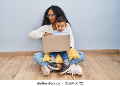 Young Hispanic Mother And Kid Using Computer Laptop Sitting On The Floor Checking The Time On Wrist Watch, Relaxed And Confident 