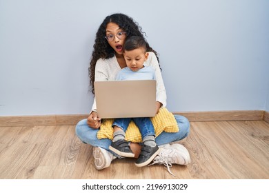 Young Hispanic Mother And Kid Using Computer Laptop Sitting On The Floor In Shock Face, Looking Skeptical And Sarcastic, Surprised With Open Mouth 