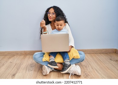 Young Hispanic Mother And Kid Using Computer Laptop Sitting On The Floor Doing Money Gesture With Hands, Asking For Salary Payment, Millionaire Business 