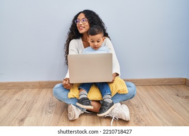 Young Hispanic Mother And Kid Using Computer Laptop Sitting On The Floor Looking Away To Side With Smile On Face, Natural Expression. Laughing Confident. 