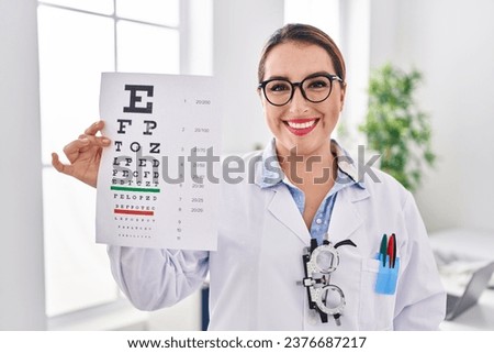 Young hispanic optician woman holding medical exam looking positive and happy standing and smiling with a confident smile showing teeth 