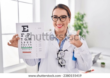 Young hispanic optician woman holding medical exam smiling happy and positive, thumb up doing excellent and approval sign 
