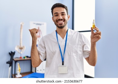 Young Hispanic Physiotherapist Man Holding Cbd Oil As Pain Relief Pointing Thumb Up To The Side Smiling Happy With Open Mouth 