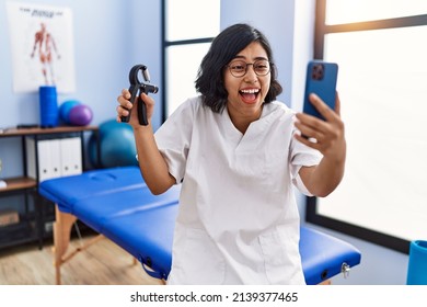 Young hispanic physiotherapist woman holding hand grip to train muscle doing video call smiling and laughing hard out loud because funny crazy joke. 