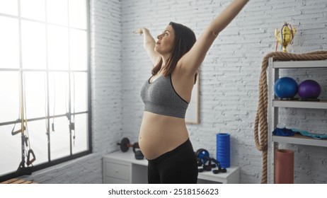 Young hispanic pregnant woman exercising in a bright indoor gym, wearing sportswear and stretching with arms raised. - Powered by Shutterstock