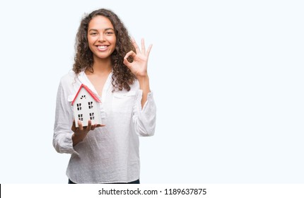 Young Hispanic Real State Agent Woman Holding Small House Doing Ok Sign With Fingers, Excellent Symbol