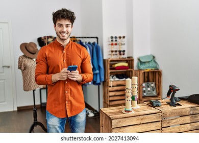 Young Hispanic Shopkeeper Man Smiling Happy Using Smartphone Working At Clothing Store.