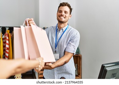 Young hispanic shopkeeper man smiling happy giving shopping bags to customer at clothing store.