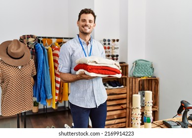 Young Hispanic Shopkeeper Man Smiling Happy Holding Stack Of Sweater At Clothing Store.