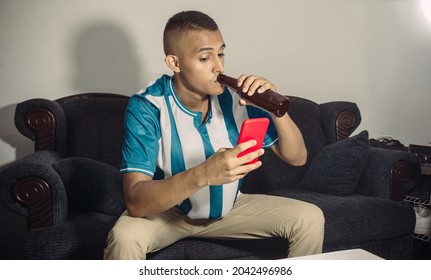 Young Hispanic Soccer Fan Man Sitting Drinking A Beer While Watching A Phone