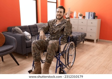 Young hispanic soldier man injured sitting on wheelchair doing ok sign with fingers, smiling friendly gesturing excellent symbol 