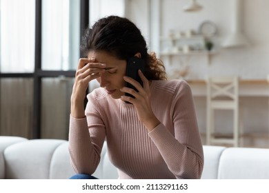 Young Hispanic stressed woman sit on sofa in living room talks on smartphone looking concerned, listen bad news feels desperate, having unpleasant remote conversation, receive disagreeable information