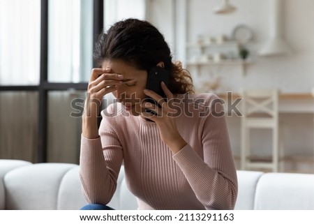Young Hispanic stressed woman sit on sofa in living room talks on smartphone looking concerned, listen bad news feels desperate, having unpleasant remote conversation, receive disagreeable information
