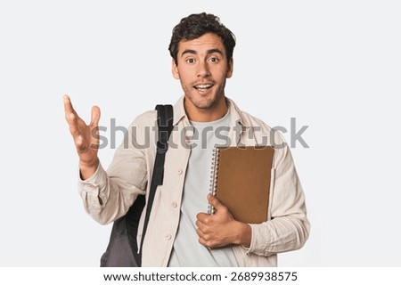 Young Hispanic student with notebook receiving a pleasant surprise, excited and raising hands.