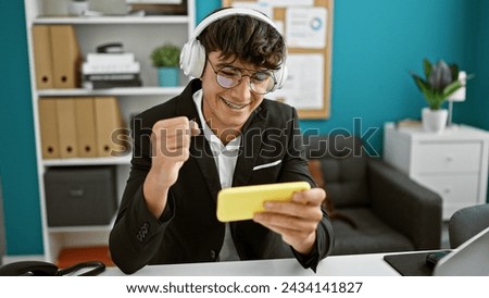 Young hispanic teenager enjoys victory in video game play at work, confidently celebrating at the office, personifying the thriving gamer business lifestyle in the indoor office background