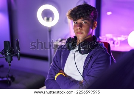 Young hispanic teenager streamer smiling confident sitting with arms crossed gesture at gaming room