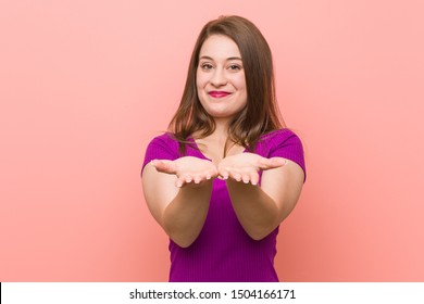 Young Hispanic Woman Against A Pink Wall Holding Something With Palms, Offering To Camera.