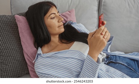 A young hispanic woman in casual attire lies on a couch in her living room, intently looking at her smartphone. - Powered by Shutterstock