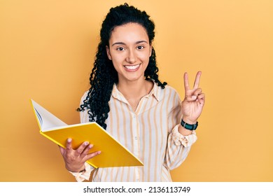 Young hispanic woman with curly hair holding book smiling looking to the camera showing fingers doing victory sign. number two. 