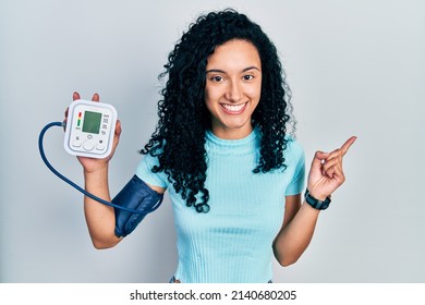 Young Hispanic Woman With Curly Hair Using Blood Pressure Monitor Smiling Happy Pointing With Hand And Finger To The Side 