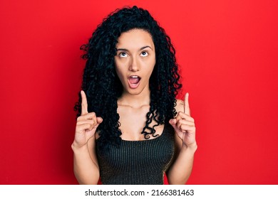 Young Hispanic Woman With Curly Hair Wearing Casual Style With Sleeveless Shirt Amazed And Surprised Looking Up And Pointing With Fingers And Raised Arms. 