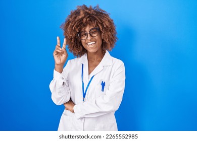 Young hispanic woman with curly hair wearing white coat and id card smiling with happy face winking at the camera doing victory sign. number two. 