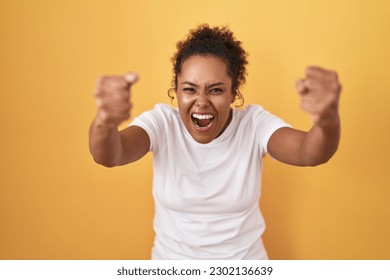 Young hispanic woman with curly hair standing over yellow background angry and mad raising fists frustrated and furious while shouting with anger. rage and aggressive concept.  - Shutterstock ID 2302136639