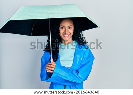 Young hispanic woman with curly hair wearing a raincoat and umbrella smiling with a happy and cool smile on face. showing teeth. 