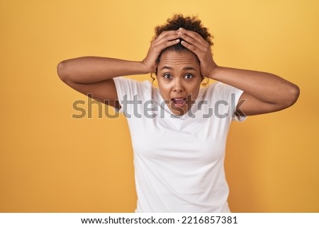 Young hispanic woman with curly hair standing over yellow background and scared with hands on head, afraid and surprised of shock with open mouth 