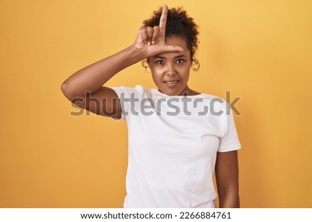 Young hispanic woman with curly hair standing over yellow background making fun of people with fingers on forehead doing loser gesture mocking and insulting. 