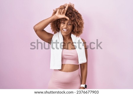 Young hispanic woman with curly hair wearing sportswear and towel making fun of people with fingers on forehead doing loser gesture mocking and insulting. 