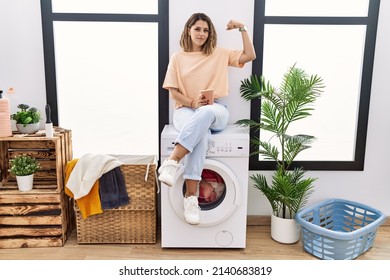 Young Hispanic Woman Drinking Coffee Waiting For Washing Machine At Laundry Room Strong Person Showing Arm Muscle, Confident And Proud Of Power 