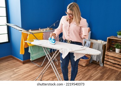 Young Hispanic Woman Drinking Coffee Ironing Clothes At Laundry Room