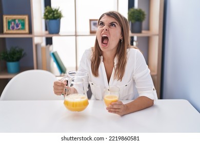 Young Hispanic Woman Drinking Glass Of Orange Juice Angry And Mad Screaming Frustrated And Furious, Shouting With Anger Looking Up. 