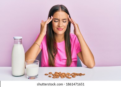 Young Hispanic Woman Drinking Healthy Almond Milk Sitting On The Table With Hand On Head, Headache Because Stress. Suffering Migraine. 