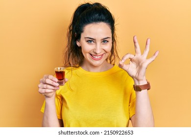 Young hispanic woman drinking whiskey shot doing ok sign with fingers, smiling friendly gesturing excellent symbol 