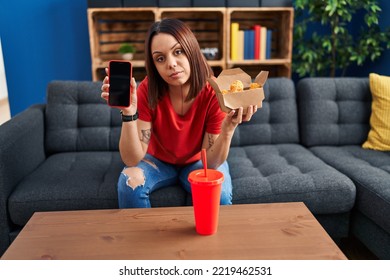 Young Hispanic Woman Eating Fast Food Showing Smartphone Screen Relaxed With Serious Expression On Face. Simple And Natural Looking At The Camera. 