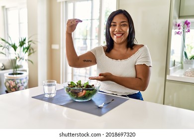 Young Hispanic Woman Eating Healthy Salad At Home Gesturing With Hands Showing Big And Large Size Sign, Measure Symbol. Smiling Looking At The Camera. Measuring Concept. 