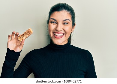 Young Hispanic Woman Eating Protein Bar As Healthy Energy Snack Celebrating Achievement With Happy Smile And Winner Expression With Raised Hand 